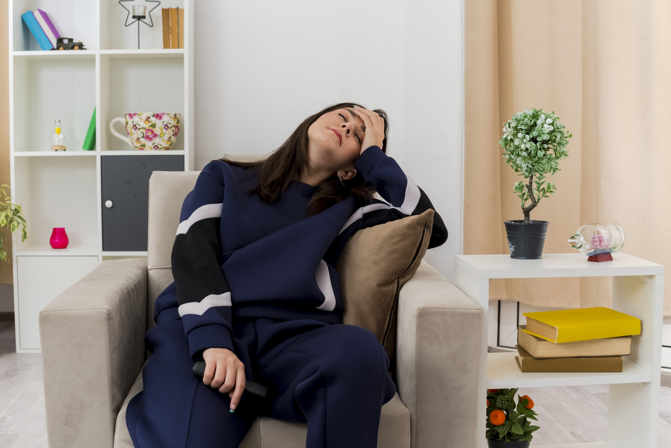 tired young pretty caucasian girl sitting on armchair in designed living room holding remote control putting hand on head with closed eyes